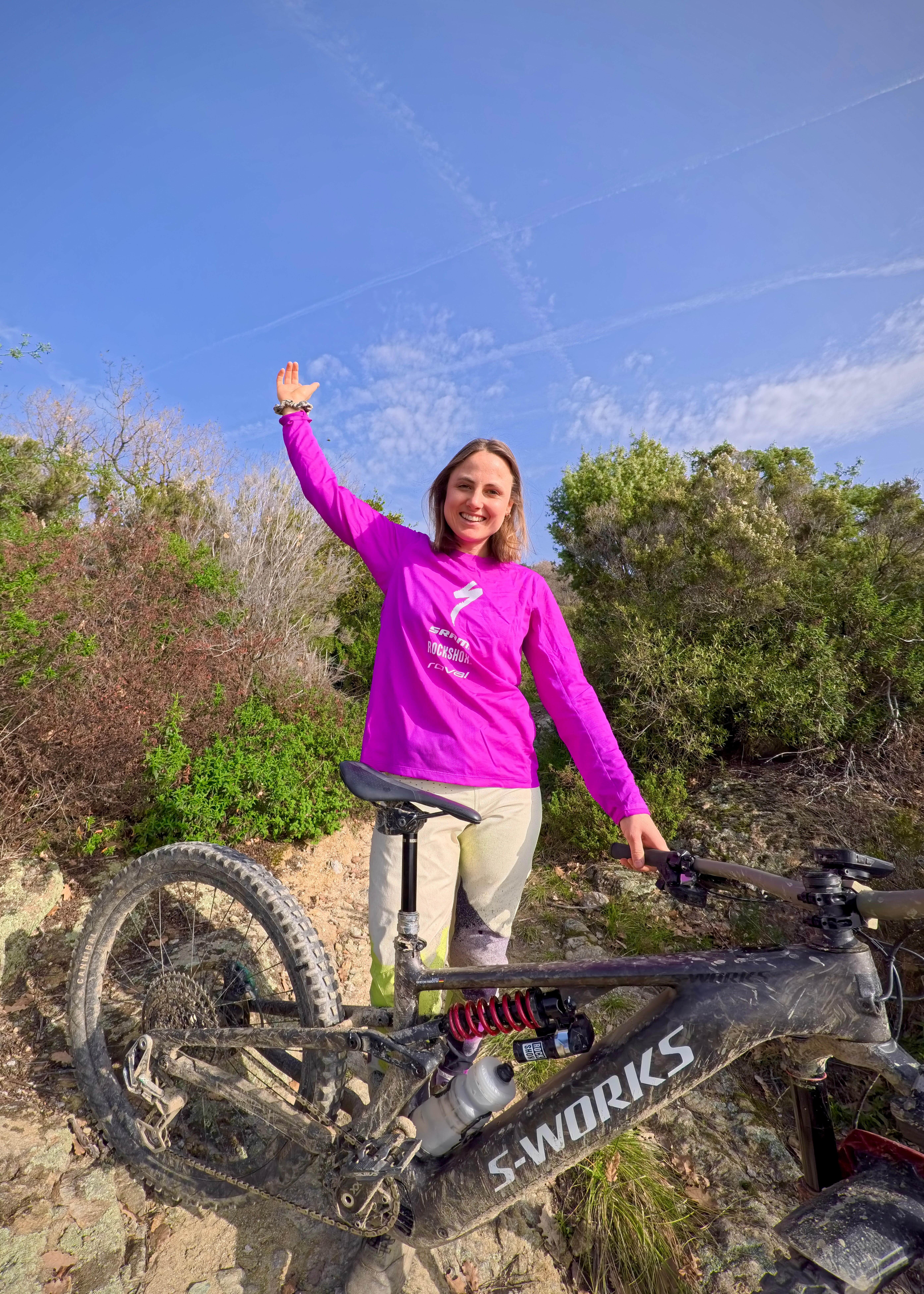 Eine lächelnde Frau im pinken Fahrradtrikot winkt neben einem schmutzigen Mountainbike auf einem steinigen Trail unter blauem Himmel.