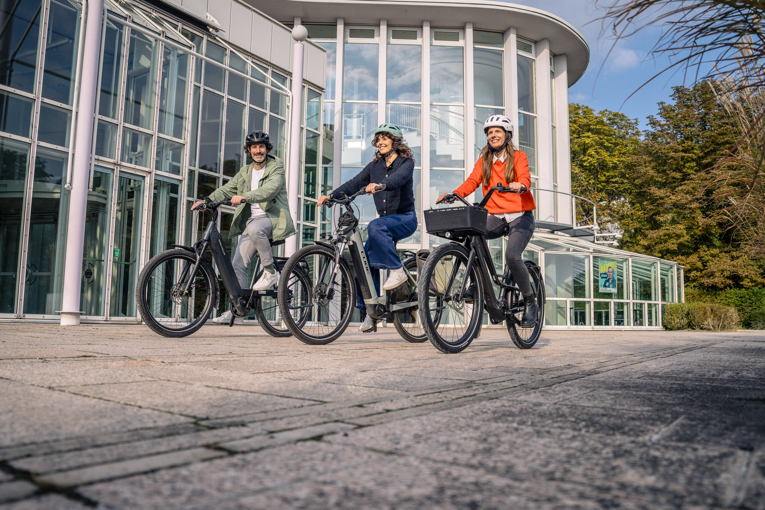 Drei lächelnde Personen fahren mit E-Bikes vor einem modernen Glasgebäude.
