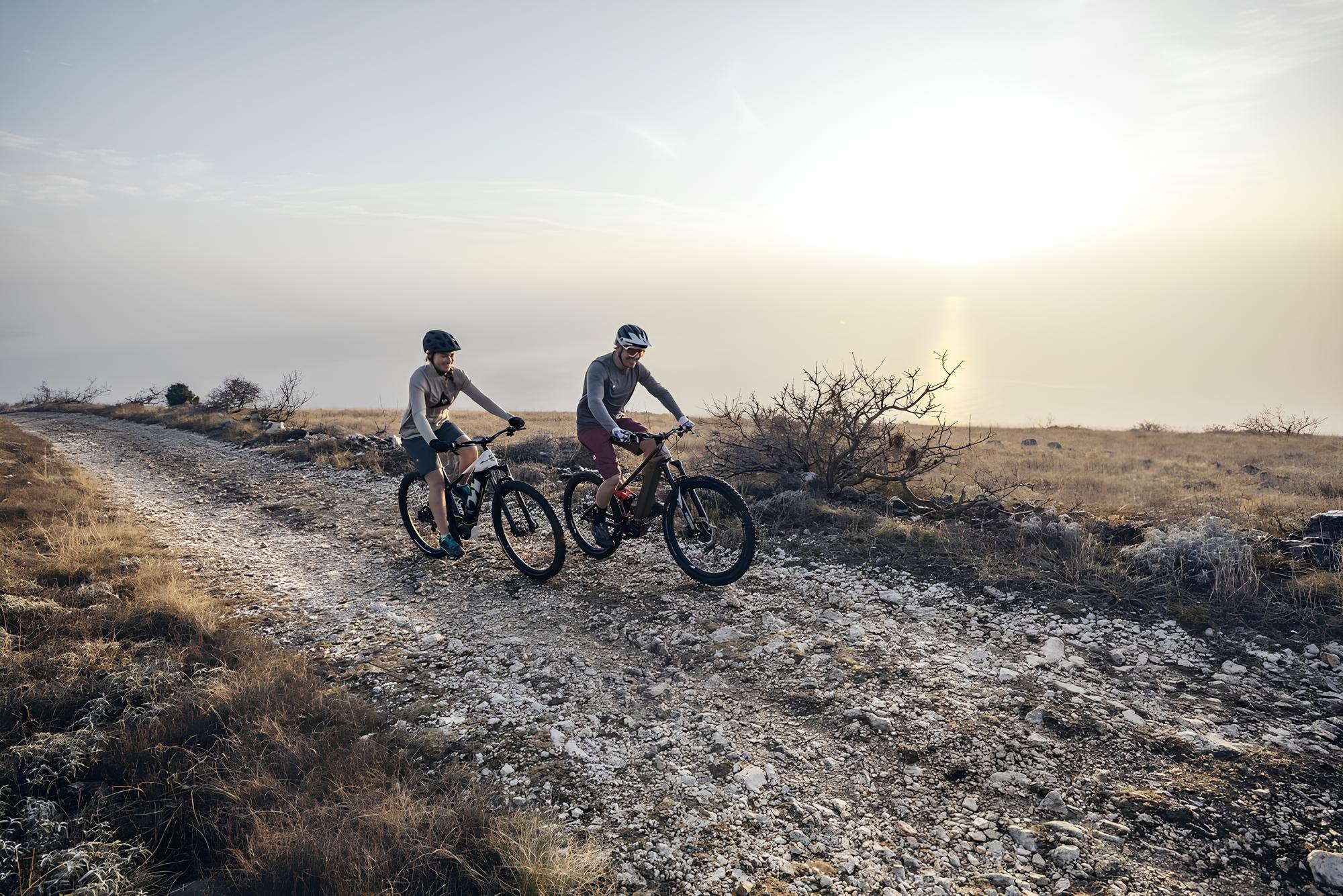 Zwei Personen fahren mit Mountainbikes auf einem steinigen Weg durch eine karge Landschaft im Sonnenlicht.