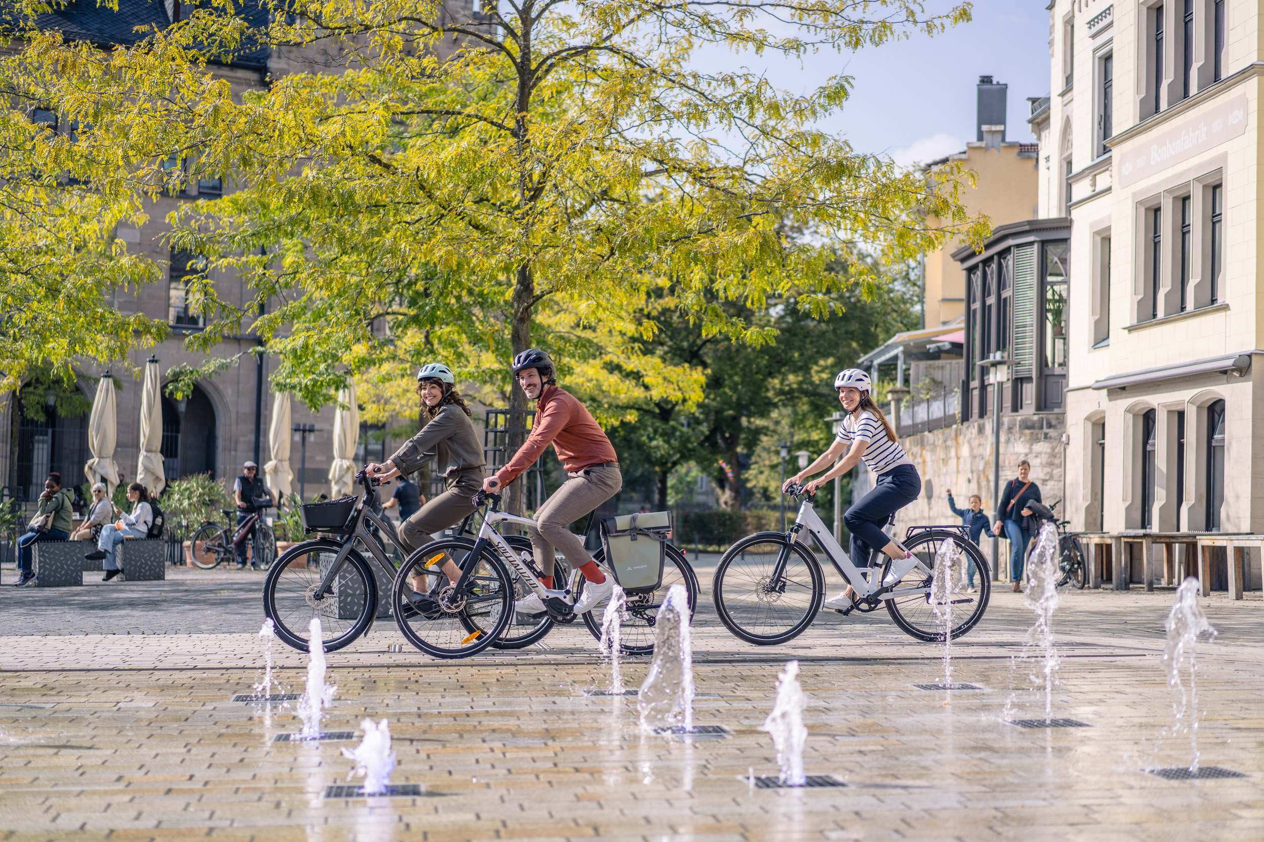 Drei Personen fahren mit E-Bikes lachend durch einen Platz mit Bodenfontänen und herbstlich gefärbten Bäumen.