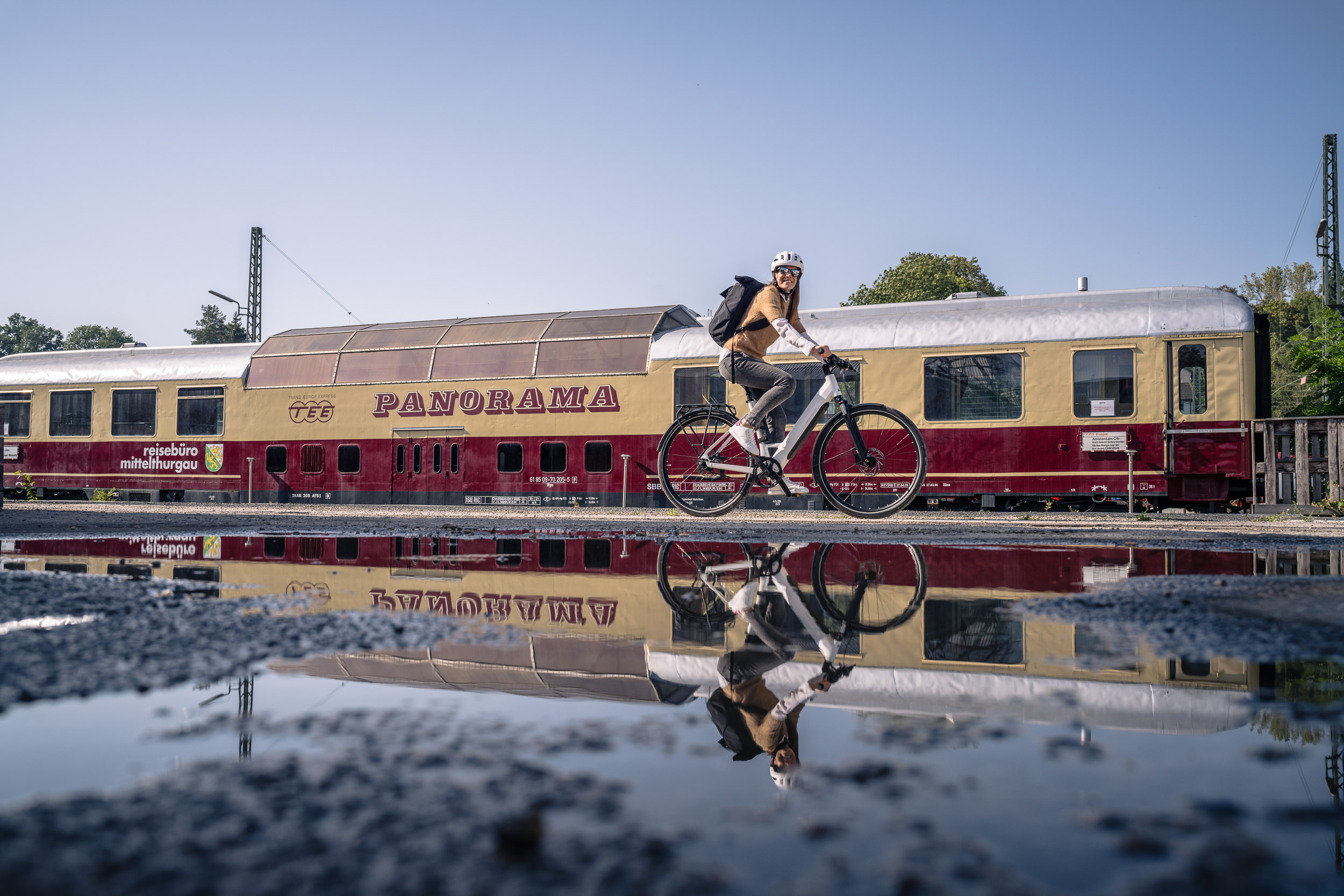 Eine Person fährt mit dem Fahrrad vor einem historischen Panoramawagen, der sich in einer Pfütze spiegelt.